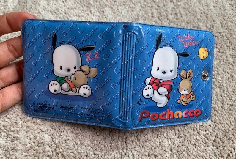 May include: A blue and white wallet with a cartoon dog and rabbit design. The wallet has the text "Pochacco" and "Waku Waku" on it.