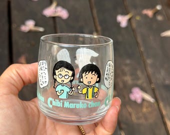 May include: A clear glass with a cartoon image of two girls from the anime "Chibi Maruko Chan". The glass has the text "Chibi Maruko Chan" printed on the bottom.
