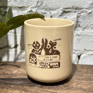 May include: A cream-colored ceramic mug with brown speckled details and a brown graphic print. The design features cartoon characters with the text "HOPPING!", "POPPING!", "LET'S SING A SONG WITH ME!", "JOLLY TIME", and "UTM SONG" printed on the front.