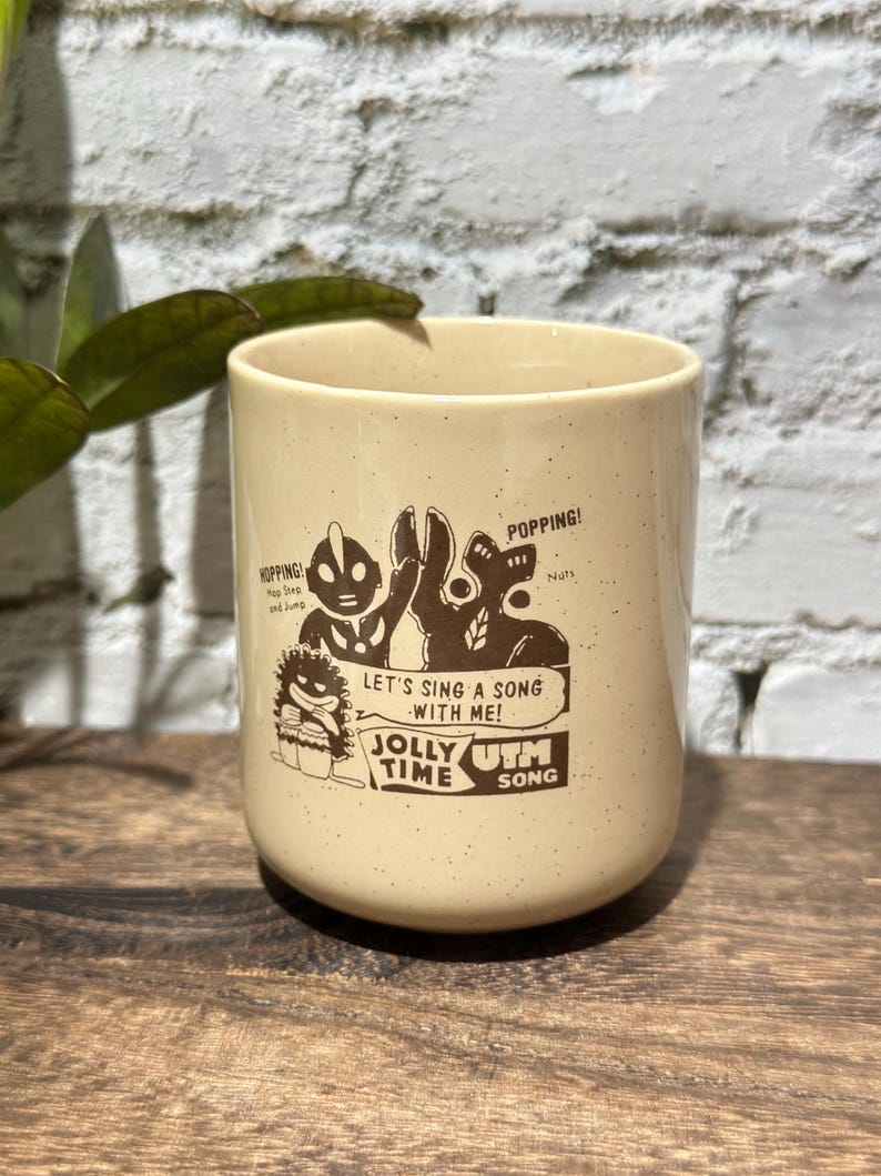 May include: A cream-colored ceramic mug with brown speckled details and a brown graphic print. The design features cartoon characters with the text "HOPPING!", "POPPING!", "LET'S SING A SONG WITH ME!", "JOLLY TIME", and "UTM SONG" printed on the front.