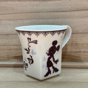 May include: White ceramic mug featuring a black silhouette of Minnie Mouse and a floral design. The mug has a square base and a handle.