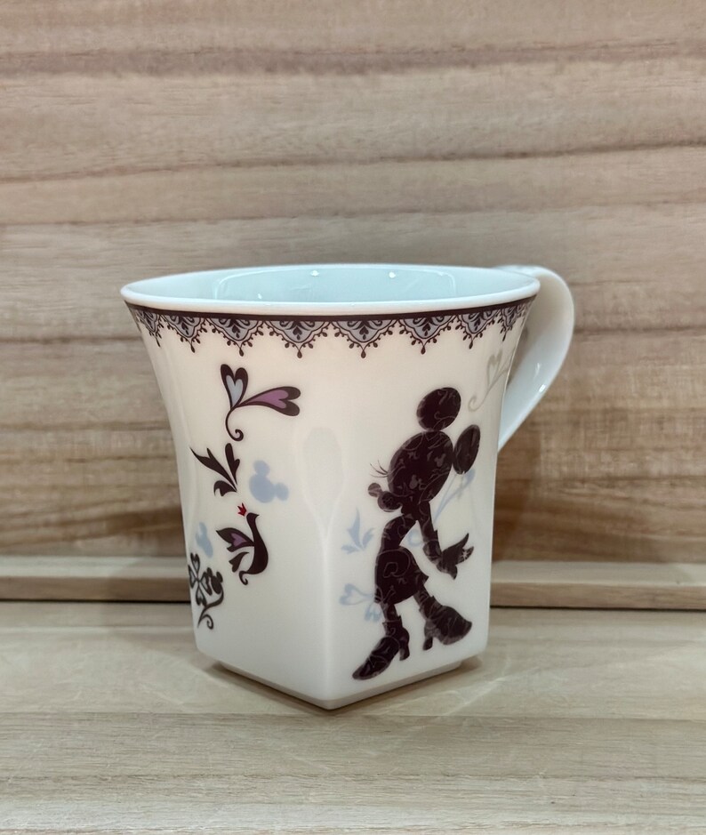 May include: White ceramic mug featuring a black silhouette of Minnie Mouse and a floral design. The mug has a square base and a handle.