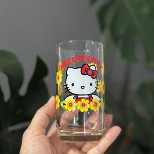 Può includere: Un bicchiere di vetro trasparente con un design di Hello Kitty. Il design presenta Hello Kitty con un vestito rosso e un fiocco rosso, circondata da fiori gialli.