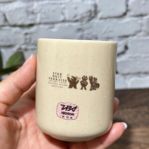 May include: A cream-colored ceramic cup with brown speckles featuring a graphic of three figures and the text STAR DUST PARADISE SINCE 1966 LIVE ACTION PROTECTOR OF THE WORLD SCIENCE FICTION, along with a circular red and white NICHION sticker.