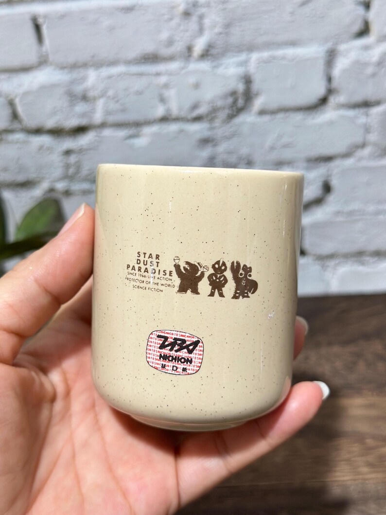May include: A cream-colored ceramic cup with brown speckles featuring a graphic of three figures and the text STAR DUST PARADISE SINCE 1966 LIVE ACTION PROTECTOR OF THE WORLD SCIENCE FICTION, along with a circular red and white NICHION sticker.