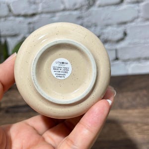 May include: A round, cream-colored ceramic object with small brown speckles. The base features a circular white sticker with black text that reads UTM 湯のみ ブラウン VICTORIA FANCY MADE IN JAPAN AK038-33 380 &copy;GAKKEN &copy;円谷プロ.