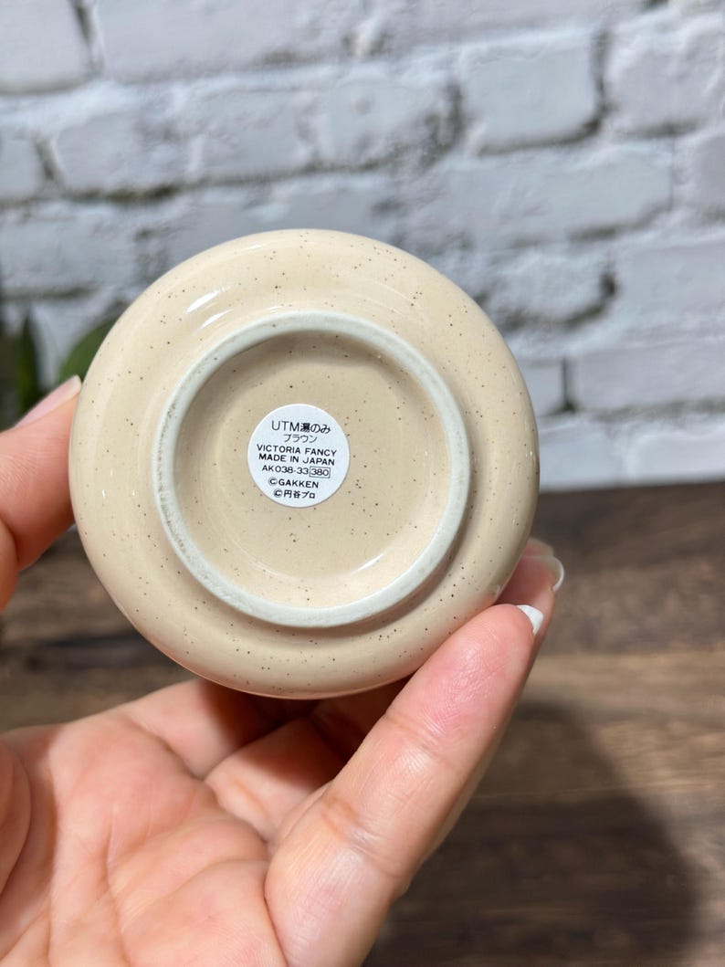 May include: A round, cream-colored ceramic object with small brown speckles. The base features a circular white sticker with black text that reads UTM 湯のみ ブラウン VICTORIA FANCY MADE IN JAPAN AK038-33 380 &copy;GAKKEN &copy;円谷プロ.