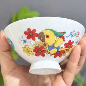 Pu&ograve; includere: Ciotola in ceramica bianca con un colorato design di pappagallo e fiori. La ciotola presenta un pappagallo giallo, blu e verde circondato da fiori rossi, gialli e bianchi. &Egrave; visibile anche del testo giapponese.