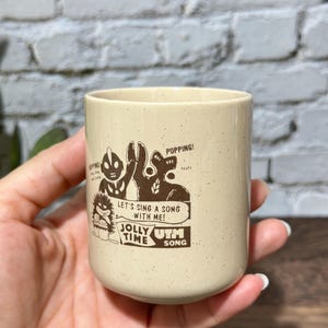 May include: A cream-colored ceramic mug with a speckled finish featuring a brown graphic print of two cartoon characters. The text reads: POPPING! LET'S SING A SONG WITH ME! JOLLY TIME UTM SONG. The mug has a cylindrical shape and a smooth rim.