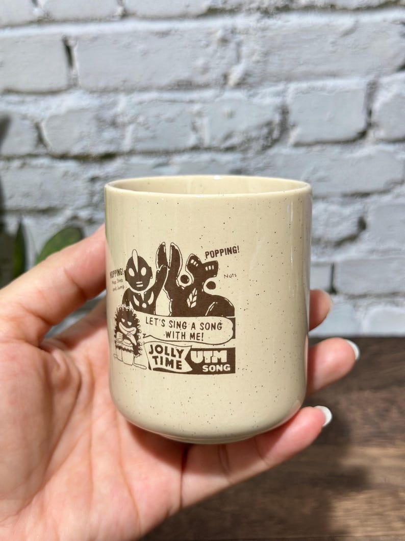May include: A cream-colored ceramic mug with a speckled finish featuring a brown graphic print of two cartoon characters. The text reads: POPPING! LET'S SING A SONG WITH ME! JOLLY TIME UTM SONG. The mug has a cylindrical shape and a smooth rim.