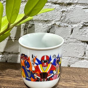 Peut inclure: Un mug en céramique blanche orné d'une impression graphique colorée représentant des personnages Power Rangers en costumes rouge, bleu et rose. Le motif montre des poses d'action avec un ranger rouge sur une moto, entouré de rangers bleu et rose.
