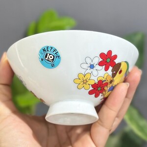 Pu&ograve; includere: Ciotola in ceramica bianca con bordo svasato e piedino. La ciotola &egrave; decorata con fiori colorati e un personaggio dei cartoni animati. Un adesivo blu con la scritta "NETTLE 10" &egrave; sul lato. La ciotola &egrave; tenuta in mano.