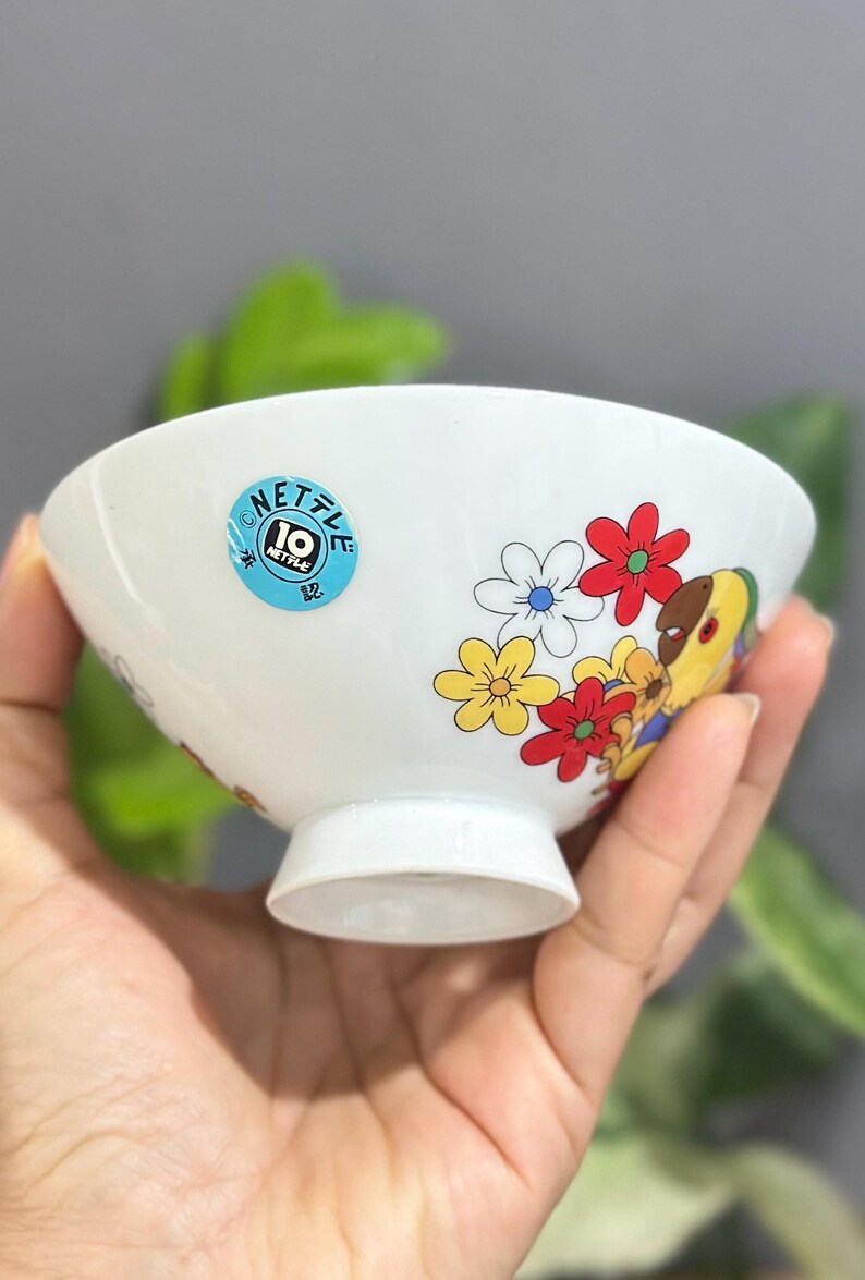 Pu&ograve; includere: Ciotola in ceramica bianca con bordo svasato e piedino. La ciotola &egrave; decorata con fiori colorati e un personaggio dei cartoni animati. Un adesivo blu con la scritta "NETTLE 10" &egrave; sul lato. La ciotola &egrave; tenuta in mano.