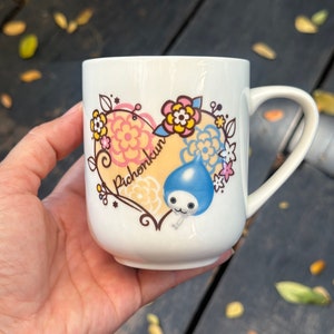 Könnte beinhalten: Eine weiße Keramiktasse mit einem herzförmigen Design, das einen blauen Comic-Charakter mit dem Text "Pichonkun" im Herzen zeigt. Das Herz ist von rosa und gelben Blumen umgeben.