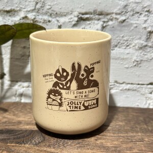 May include: A cream-colored ceramic mug with a speckled finish featuring a brown graphic print of Ultraman characters. The text reads: HOPPING! Hop Step and Jump, POPPING! Nuts, LET'S SING A SONG WITH ME! JOLLY TIME UTM SONG.