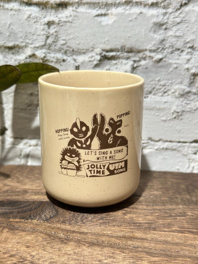 May include: A cream-colored ceramic mug with a speckled finish featuring a brown graphic print of Ultraman characters. The text reads: HOPPING! Hop Step and Jump, POPPING! Nuts, LET'S SING A SONG WITH ME! JOLLY TIME UTM SONG.