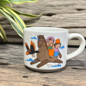 Peut inclure: Mug en céramique blanche avec une illustration de dessin animé de deux enfants montant un cheval marron, avec un oiseau noir volant à proximité. La tasse a une anse incurvée et le texte "日本アニメ" en bas.