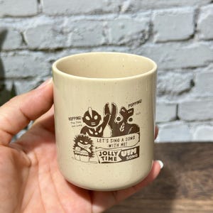 May include: A cream-colored ceramic mug with a speckled finish featuring a brown graphic print of two characters. The text reads: HOPPING! Hop Step and Jump, POPPING!, LET'S SING A SONG WITH ME!, JOLLY TIME, and UTM SONG.
