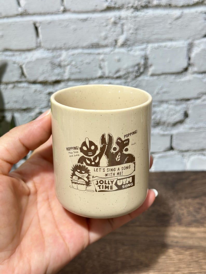 May include: A cream-colored ceramic mug with a speckled finish featuring a brown graphic print of two characters. The text reads: HOPPING! Hop Step and Jump, POPPING!, LET'S SING A SONG WITH ME!, JOLLY TIME, and UTM SONG.