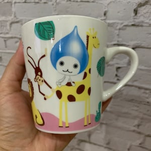 May include: White ceramic mug with a colourful cartoon design featuring a giraffe, monkey, and a blue raindrop character. The mug has a pink rim and a brown handle.