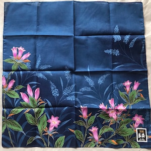Pode incluir: Um lenço de algodão azul com um padrão floral. As flores são rosa e brancas com folhas verdes. O lenço tem uma borda dourada e um pequeno rótulo com texto japonês.
