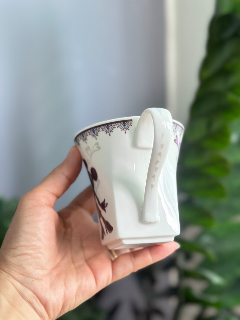 May include: White ceramic teacup with a brown and white floral design and a handle with small heart details.
