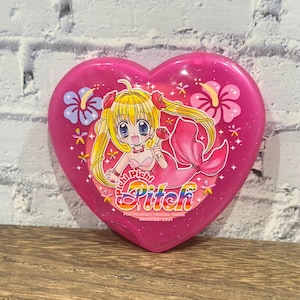 Puede incluir: Un estuche compacto rosa en forma de corazón con una sirena de dibujos animados de pelo rubio y cola rosa. El estuche tiene un acabado brillante y está decorado con motivos florales y el texto "Pichi Pichi Pitch".