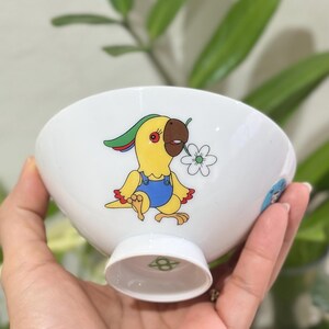 Pu&ograve; includere: Ciotola in ceramica bianca con un disegno di pappagallo dei cartoni animati. Il pappagallo &egrave; giallo con becco marrone, indossa una tuta blu e tiene in mano un fiore bianco. La ciotola ha un piedino e un logo verde e bianco sul fondo.