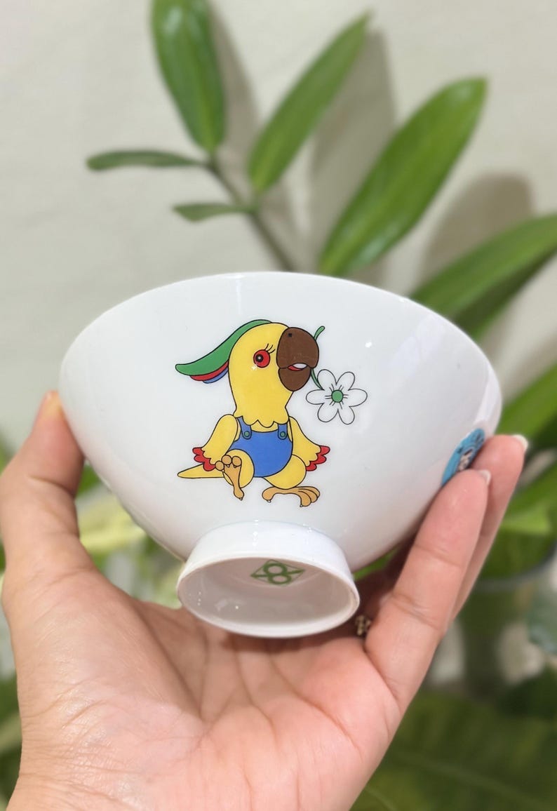 Pu&ograve; includere: Ciotola in ceramica bianca con un disegno di pappagallo dei cartoni animati. Il pappagallo &egrave; giallo con becco marrone, indossa una tuta blu e tiene in mano un fiore bianco. La ciotola ha un piedino e un logo verde e bianco sul fondo.