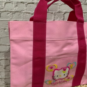 K&ouml;nnte beinhalten: Rosa Hello Kitty-Tragetasche mit roten Tr&auml;gern und einem Hello Kitty-Motiv mit Blumen.