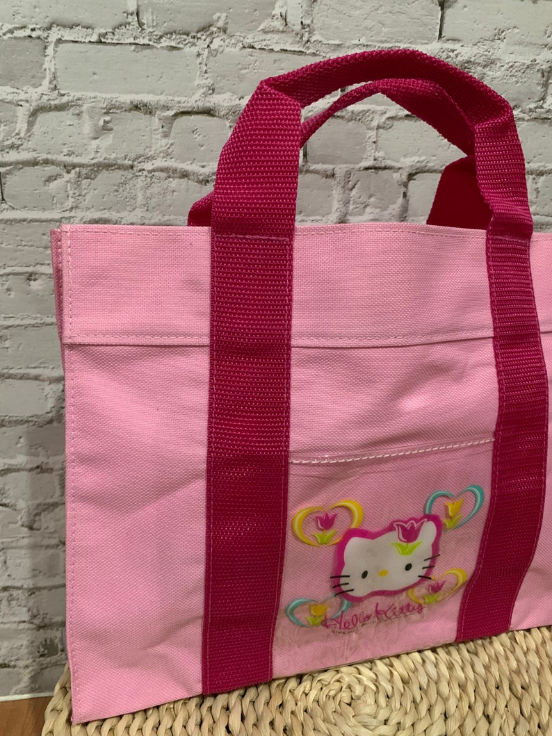 K&ouml;nnte beinhalten: Rosa Hello Kitty-Tragetasche mit roten Tr&auml;gern und einem Hello Kitty-Motiv mit Blumen.
