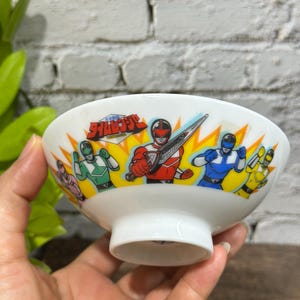 May include: A white ceramic bowl featuring a colorful graphic print of the Power Rangers Time Force characters in red, blue, green, yellow, and pink suits. The design includes a stylized logo with Japanese text and yellow lightning bolt accents.