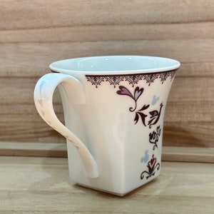 May include: White ceramic mug with a square base and a handle. The mug features a floral design with a bird and hearts in shades of pink, purple, and blue.