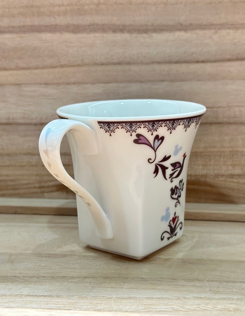 May include: White ceramic mug with a square base and a handle. The mug features a floral design with a bird and hearts in shades of pink, purple, and blue.