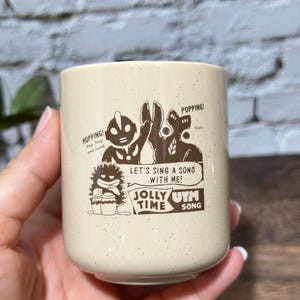 May include: A cream-colored ceramic mug with brown speckled details and a brown graphic print. The design features cartoon characters, the text "HOPPING!", "POPPING!", "LET'S SING A SONG WITH ME!", "JOLLY TIME", and "UTM SONG".