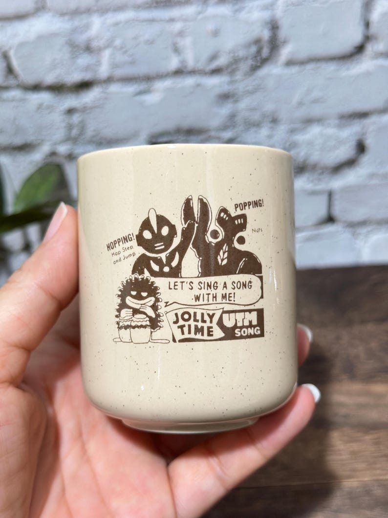 May include: A cream-colored ceramic mug with brown speckled details and a brown graphic print. The design features cartoon characters, the text "HOPPING!", "POPPING!", "LET'S SING A SONG WITH ME!", "JOLLY TIME", and "UTM SONG".