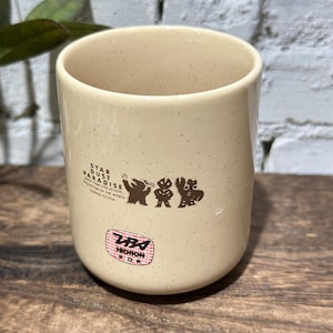 May include: A cream-colored ceramic mug with a speckled finish. The front features brown graphic silhouettes of three figures and the text STAR DUST PARADISE, along with a circular red and white sticker containing the text Tsuburaya and Nichion.