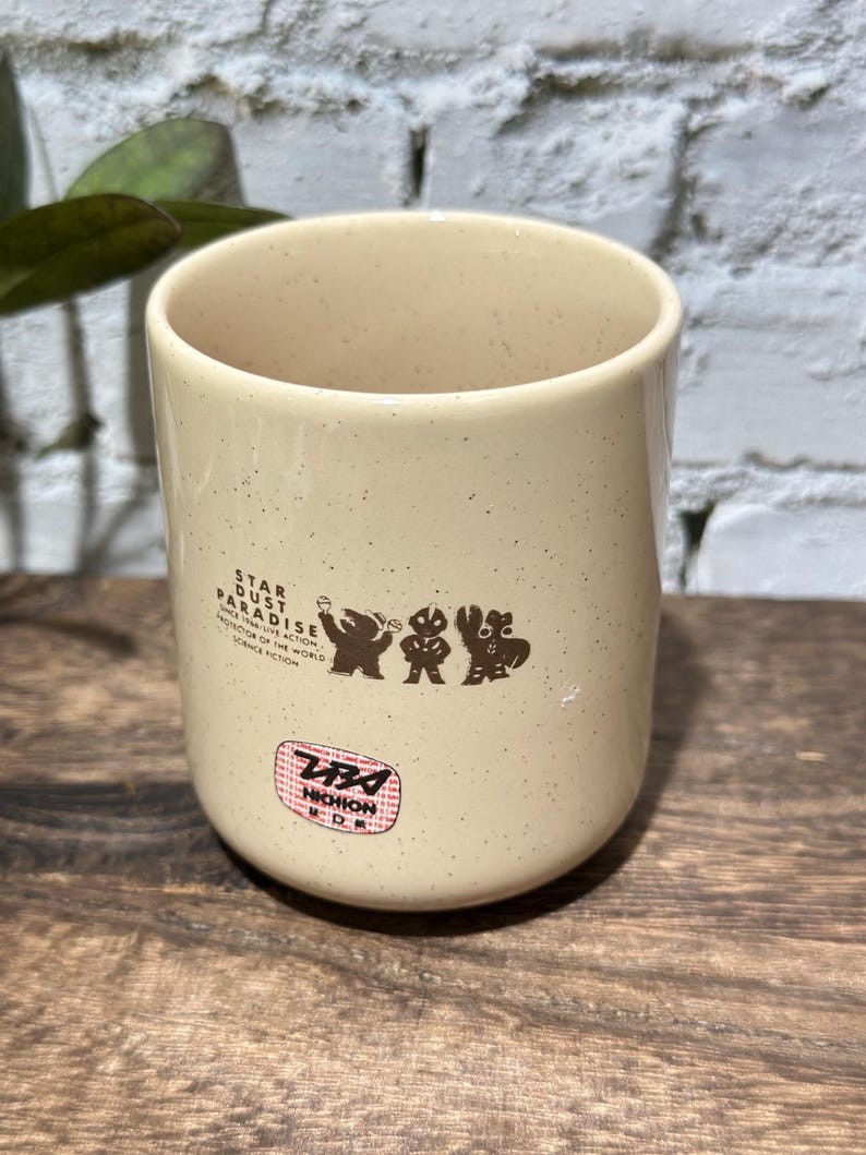 May include: A cream-colored ceramic mug with a speckled finish. The front features brown graphic silhouettes of three figures and the text STAR DUST PARADISE, along with a circular red and white sticker containing the text Tsuburaya and Nichion.