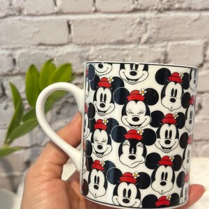 Puede incluir: Taza de cer&aacute;mica blanca con un patr&oacute;n repetido de caras de Mickey Mouse con orejas negras, un sombrero rojo y una expresi&oacute;n sonriente. La taza tiene un asa blanca y un borde negro.