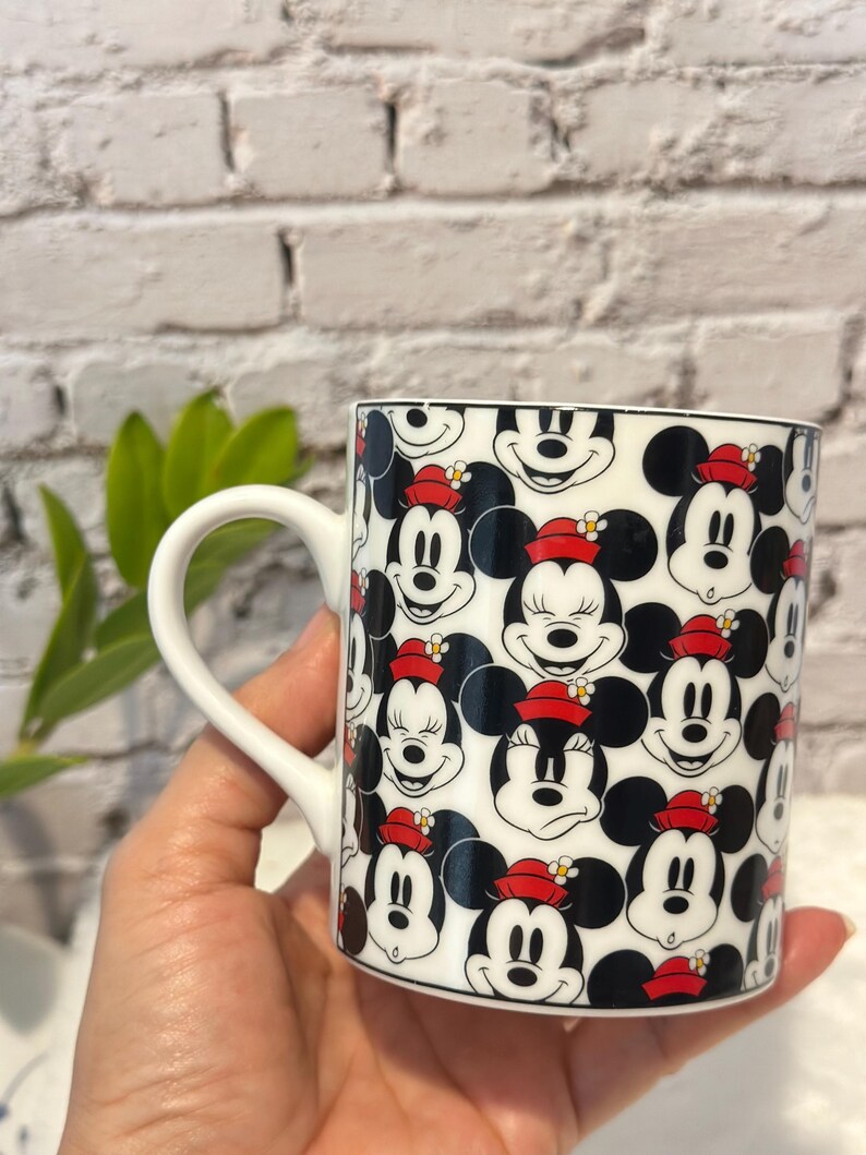 Puede incluir: Taza de cer&aacute;mica blanca con un patr&oacute;n repetido de caras de Mickey Mouse con orejas negras, un sombrero rojo y una expresi&oacute;n sonriente. La taza tiene un asa blanca y un borde negro.