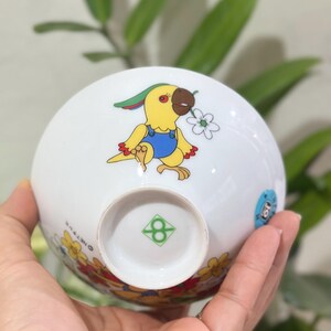 Pu&ograve; includere: Ciotola in ceramica bianca con un pappagallo dei cartoni animati in tuta gialla che tiene un fiore. La ciotola ha un bordo floreale e un logo verde sul fondo. La ciotola &egrave; tenuta in una mano.