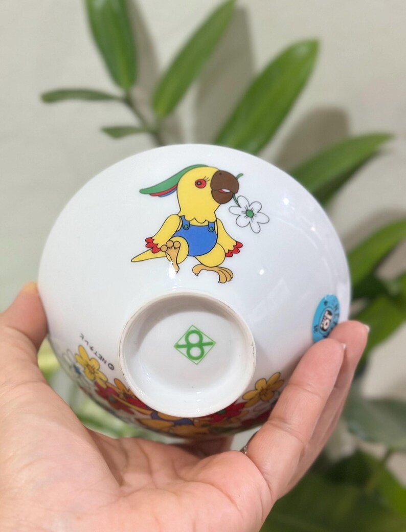 Pu&ograve; includere: Ciotola in ceramica bianca con un pappagallo dei cartoni animati in tuta gialla che tiene un fiore. La ciotola ha un bordo floreale e un logo verde sul fondo. La ciotola &egrave; tenuta in una mano.