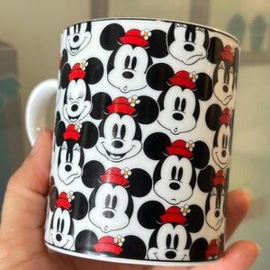 Puede incluir: Una taza de cer&aacute;mica blanca con un patr&oacute;n repetido de caras de Mickey Mouse. Las caras son en blanco y negro con sombreros rojos y peque&ntilde;os detalles florales. La taza tiene un asa blanca y un borde negro.