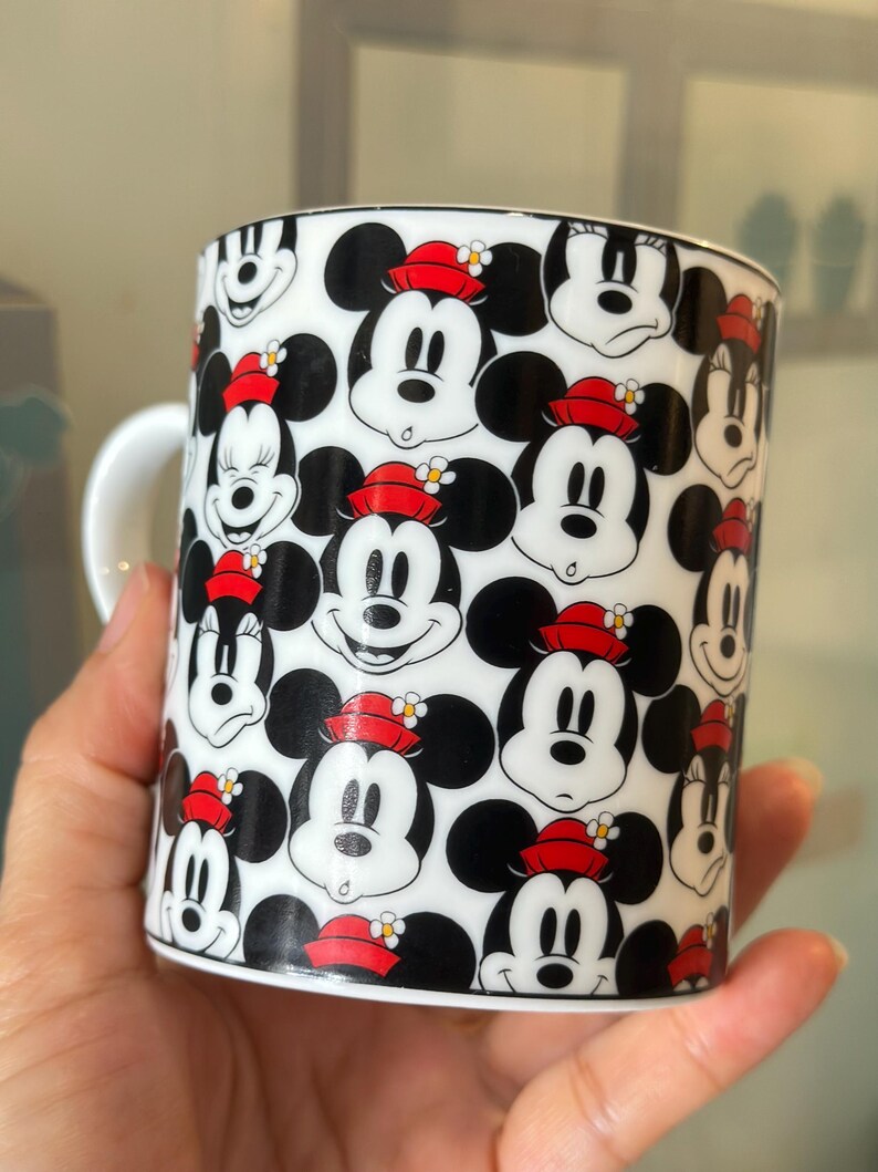 Puede incluir: Una taza de cer&aacute;mica blanca con un patr&oacute;n repetido de caras de Mickey Mouse. Las caras son en blanco y negro con sombreros rojos y peque&ntilde;os detalles florales. La taza tiene un asa blanca y un borde negro.