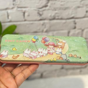 May include: A rectangular pencil case with a pink border and a green top featuring a cartoon scene. The scene includes rabbits, a girl in a hat, balloons, and the text "TINY CANDY".