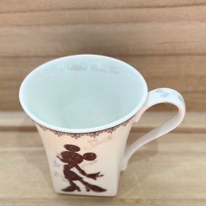 May include: White ceramic mug featuring a brown silhouette of Mickey Mouse on the front. The mug has a white lace design around the top and a white handle.
