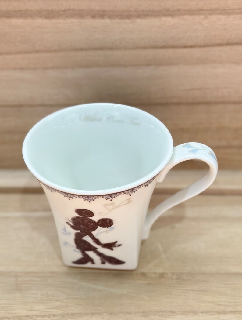 May include: White ceramic mug featuring a brown silhouette of Mickey Mouse on the front. The mug has a white lace design around the top and a white handle.