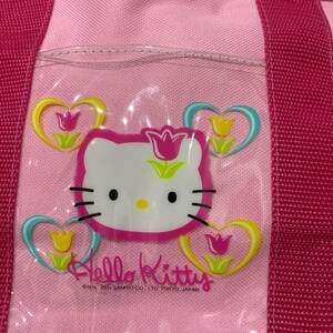 K&ouml;nnte beinhalten: Rosa Hello Kitty-Tragetasche mit einem durchsichtigen Plastikfenster, das Hello Kitty und ein Blumenmuster zeigt. Der Text "Hello Kitty" und "&copy;1976, 2001 SANRIO CO., LTD. TOKYO, JAPAN" ist auf der Tasche aufgedruckt.