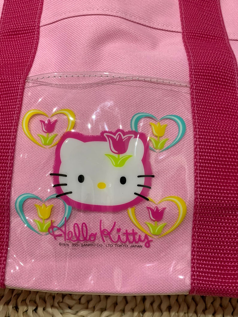 K&ouml;nnte beinhalten: Rosa Hello Kitty-Tragetasche mit einem durchsichtigen Plastikfenster, das Hello Kitty und ein Blumenmuster zeigt. Der Text "Hello Kitty" und "&copy;1976, 2001 SANRIO CO., LTD. TOKYO, JAPAN" ist auf der Tasche aufgedruckt.