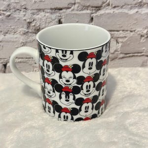 Puede incluir: Taza de cer&aacute;mica blanca con un patr&oacute;n repetido de caras de Mickey Mouse con sombreros rojos. La taza tiene un asa blanca y un borde negro. El fondo es una pared de ladrillos blancos y una superficie blanca y esponjosa.